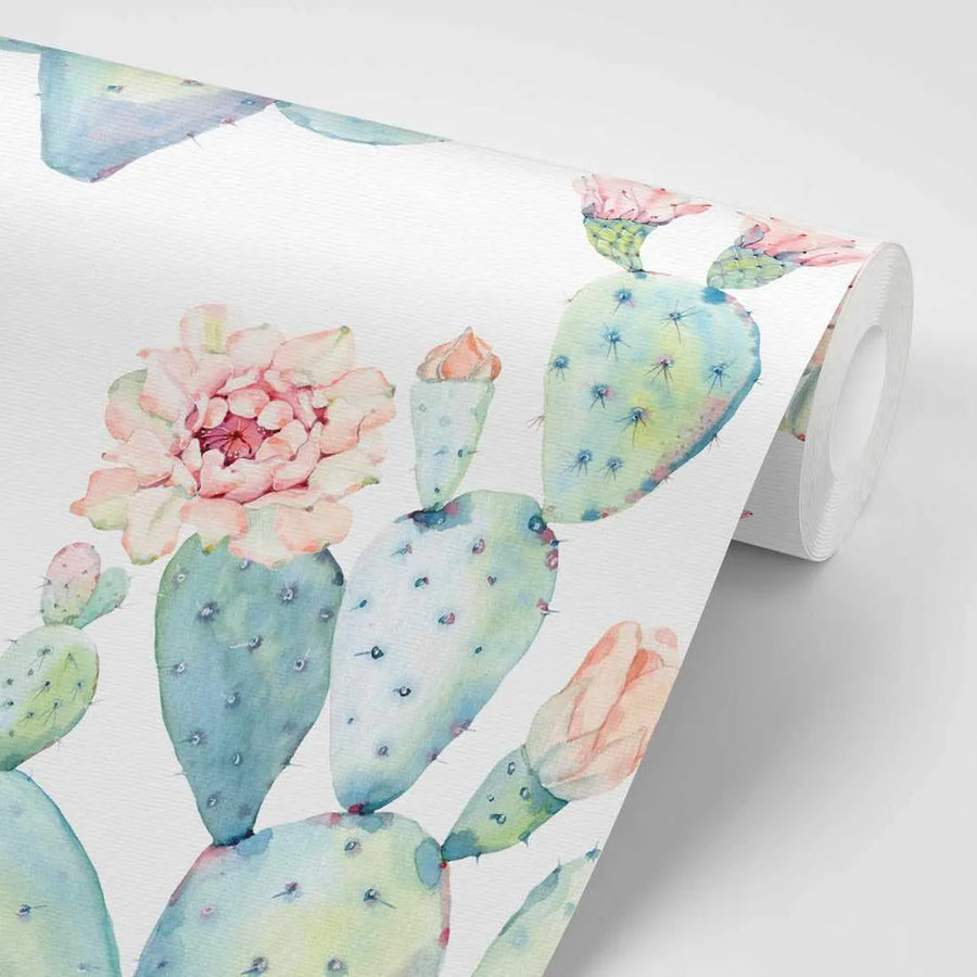 "Peel and stick wallpaper featuring a repeating pattern of hand-painted prickly pear cacti in various shades of blue-green, sage, and pale teal, with some pads adorned with soft pink and peach blossoms. The watercolour design has a loose, organic quality with visible brushstroke texture, and the wallpaper is shown partially unrolled against a white background."
