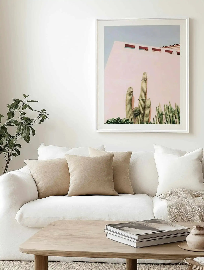 Framed art print displayed above a cream upholstered sofa in a minimalist living room. The photograph shows a tall saguaro cactus with smaller succulents against a dusty rose pink wall of a southwestern adobe building with small rectangular windows and terracotta roof tiles visible at top. Clear blue sky above. White wooden frame with white border. Natural light wood coffee table with stacked magazines below. Potted green plant visible to the left.