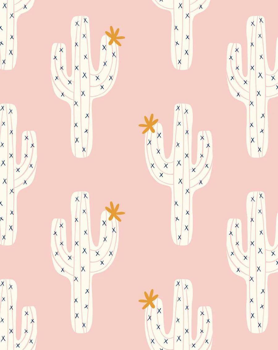 Peel and stick wallpaper featuring a repeating pattern of hand-drawn saguaro cacti in off-white with small navy blue 'x' marks representing needles, on a soft blush pink background. Some cacti have a small, five-petal orange flower blooming from their arms or tops. The illustration style is simple and playful.