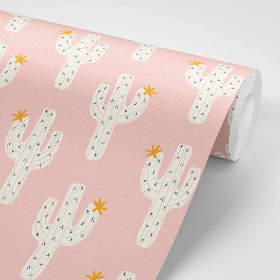 Peel and stick wallpaper featuring a repeating pattern of hand-drawn saguaro cacti in off-white with small navy blue 'x' marks for spines and a yellow-orange star-shaped flower on top, set against a soft blush pink background. The design has a playful, illustrative quality.