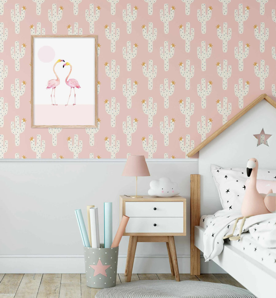 Pink wallpaper with a repeating pattern of white saguaro cacti with small yellow flowers. The wallpaper is displayed in a child's bedroom with a white bed frame, a light wood bedside table, and a framed print of two pink flamingos.
