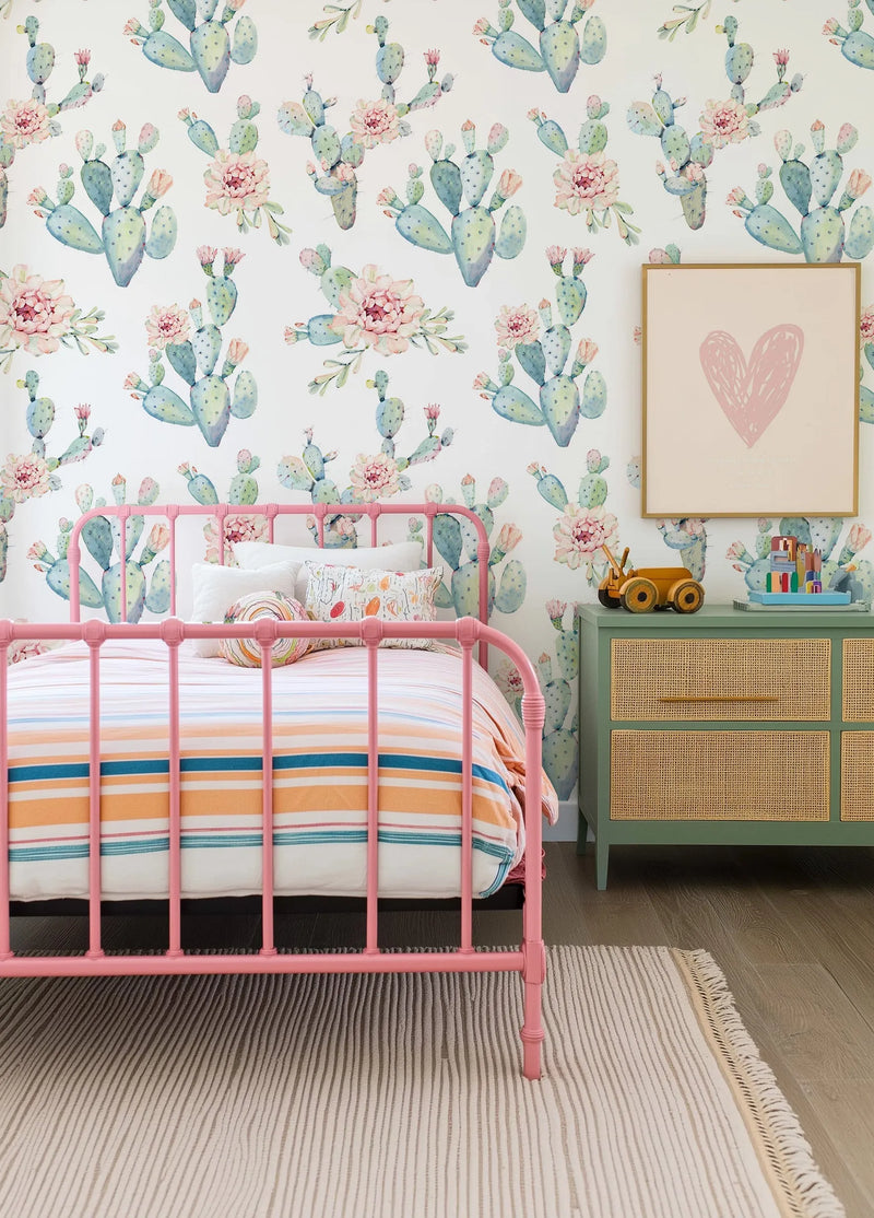 "A child's bedroom featuring a pink metal bed frame with a striped duvet and a mint green dresser with rattan drawer fronts. The walls are covered in a wallpaper featuring a repeating pattern of watercolor prickly pear cacti in shades of blue-green and sage, with soft pink flowers and buds on a white background. Above the dresser hangs a framed art print with a pink heart illustration."