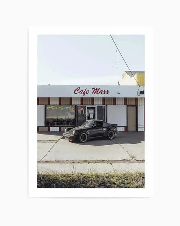 Framed photograph of a vintage black Porsche parked in front of an old-fashioned diner called "Cafe Maxx" under a bright, clear sky. The cafe has a white facade with red stripes and red lettering, and its windows display signs for "BURGERS," "FISH & CHIPS," "SALAD ROLLS," and "COFFEE." The car is dark-colored with black wheels, and the foreground shows a concrete lot with patches of dry grass and weeds.