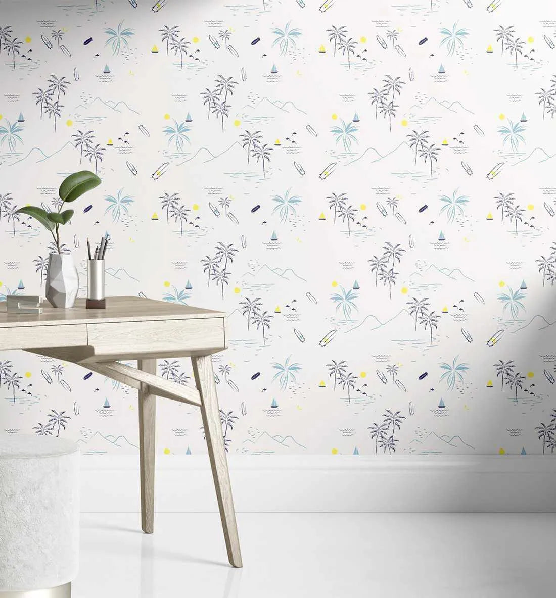 Lifestyle shot of a home office featuring a light wood desk and a white stool against a wall covered in Cali' Dreams wallpaper. The wallpaper features a repeating pattern of hand-drawn palm trees, mountains, ocean waves, sailboats, surfboards, and birds in a playful, minimalist style with a white background. The design uses a palette of navy blue, light blue, yellow, and black.