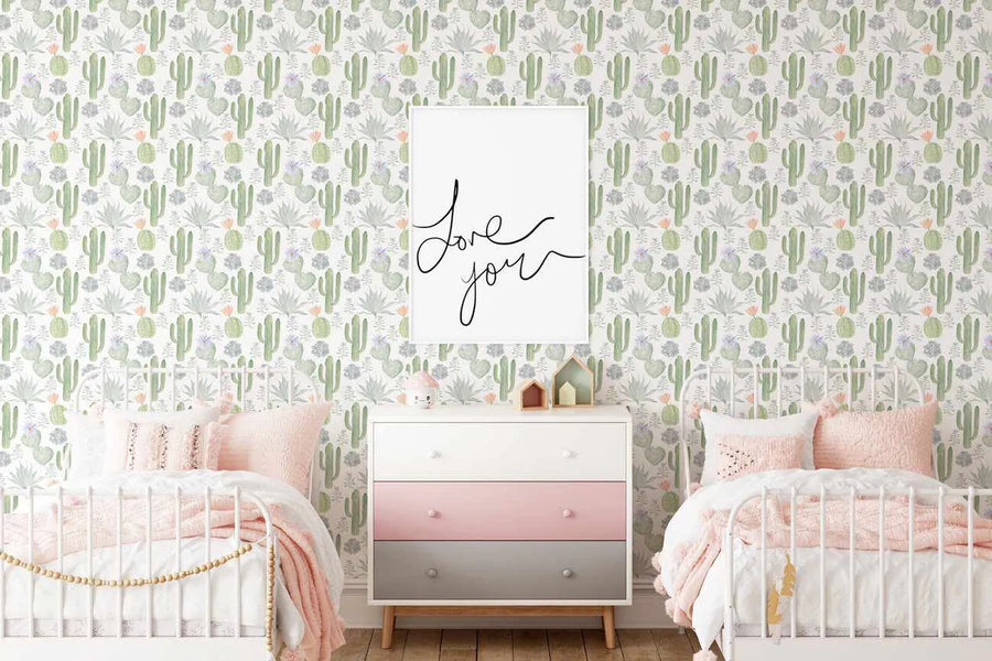 Two white metal-framed twin beds with pink bedding and pillows flank a white dresser with pink and grey drawers, all set against a wallpaper featuring a repeating pattern of hand-painted cacti and succulents in various shades of green, grey, and subtle orange and purple blooms on a white background. A framed print with the words "Love you" in black script hangs above the dresser.