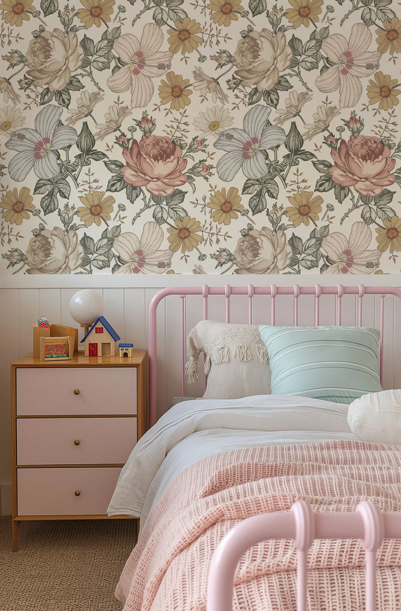 "A child's bedroom with a pink metal bed frame and a light wood bedside table with two drawers. The wall behind the bed is covered in wallpaper featuring a repeating pattern of large-scale floral illustrations in muted tones of dusty rose, cream, pale blue, and mustard yellow against a light cream background. The bed is dressed with white linen, a light pink textured throw, and pillows in cream and pale blue stripes. The bedside table holds a white globe lamp and small wooden house toys."