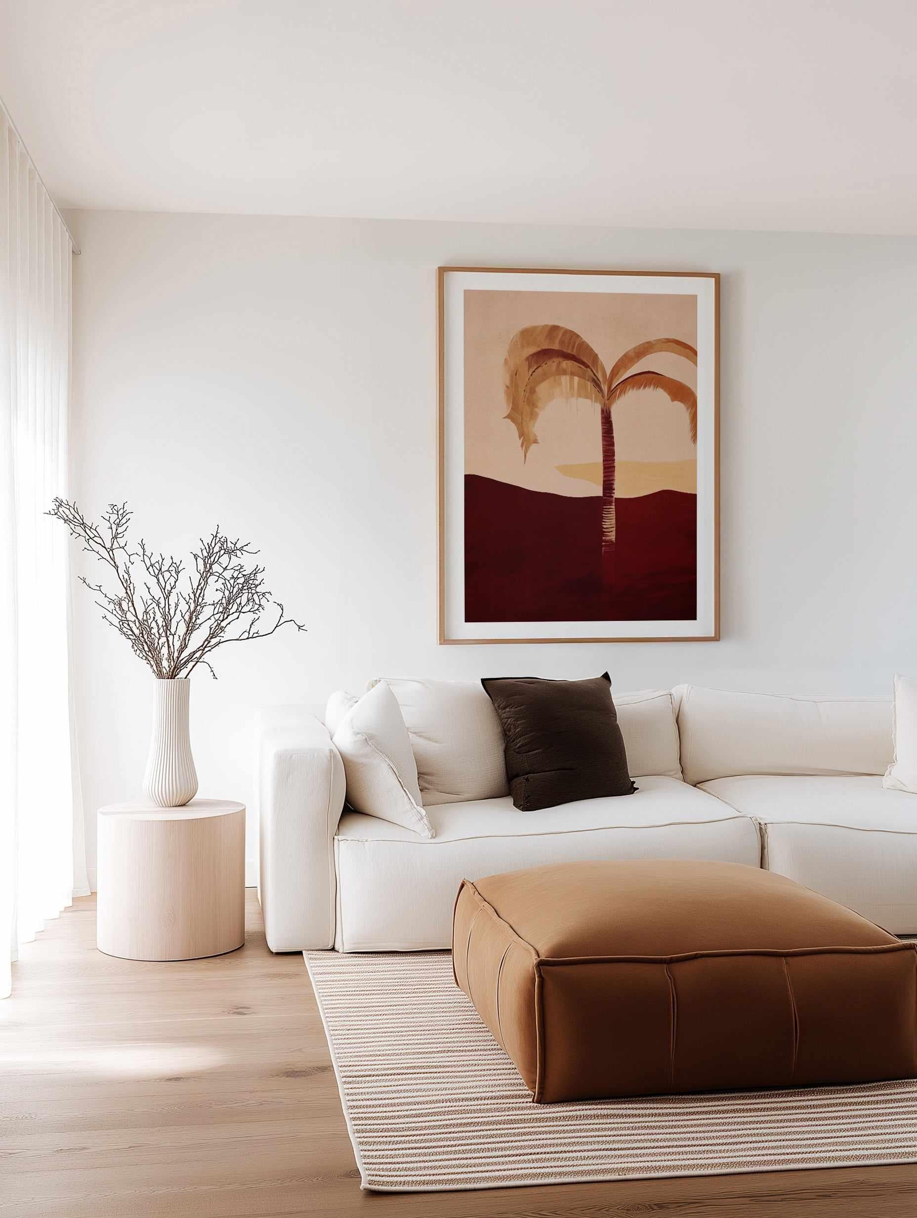 Modern living room with a white sofa, brown ottoman, and framed burgundy toned palm tree wall print artwork on the wall.