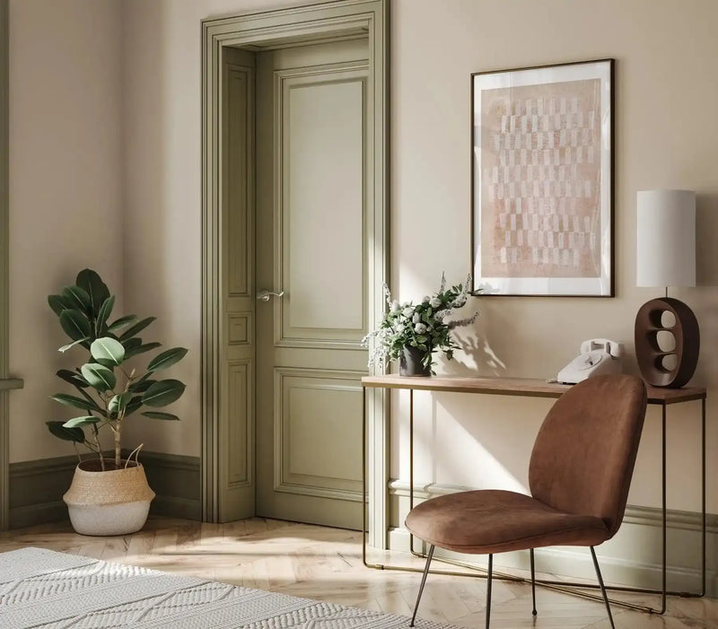 Lifestyle shot of a framed art print displayed above a wooden console table in a neutral-toned hallway. The print features an abstract pattern of small, irregular rectangular shapes in white and muted pinkish-brown, arranged in a grid-like fashion against a light beige background, with a dark brown frame and white border. The room includes a large green plant in a woven basket, a white rotary phone, a dark brown sculptural lamp with a white shade, and a brown upholstered chair.