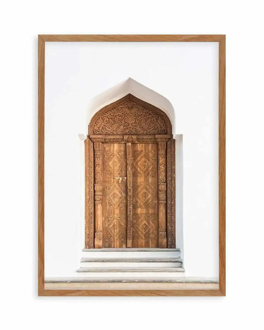 Framed photography art print of an intricately carved wooden door with a natural oak frame and white border. The door features a pointed arch at the top with elaborate floral and vine carvings, and two vertical panels with a repeating diamond pattern. Three light grey steps lead up to the door, which is set into a plain white wall.