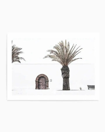 Black and white photograph art print featuring a minimalist outdoor scene with a white wall, an arched wooden door, a tall palm tree, a small shrub, and a wooden bench. The composition is stark and high-contrast, with the dark elements standing out against the bright white background.