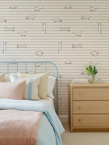 "Bedroom featuring wallpaper with a repeating pattern of continuous line art cats in various poses on a cream background with thin horizontal stripes. The wallpaper is displayed behind a light blue metal bed frame with a bed dressed in light blue and pink bedding, and a light wood bedside table with a vase of greenery and books."