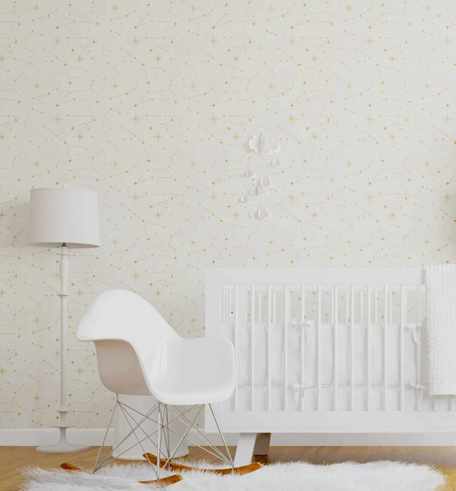 Nursery:
Celestial Night Sky wallpaper featuring a repeating pattern of hand-drawn constellations and scattered stars in soft gold and pale yellow on a creamy white background. The wallpaper is displayed in a minimalist nursery with a white crib, a white rocking chair, a white floor lamp, and a white fluffy rug. A white cloud mobile hangs above the crib.