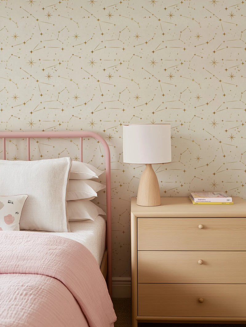 "Celestial Night Sky Wallpaper featuring a repeating pattern of gold constellations and scattered stars on a light cream background, displayed on a bedroom wall behind a pink metal bed frame. The bed is dressed with white pillows and a dusty pink textured duvet, with a light wood bedside table and lamp to the right."