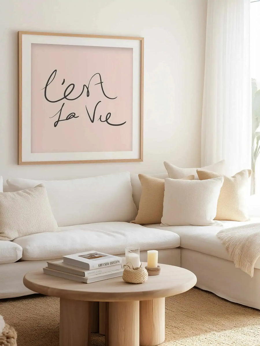 Square art print with blush pink background featuring handwritten text reading "C'est la Vie" in dark gray script, displayed in light natural wood frame with white border, hung above a cream curved sofa styled with neutral pillows in a minimalist living room with wooden coffee table and natural lighting from window.