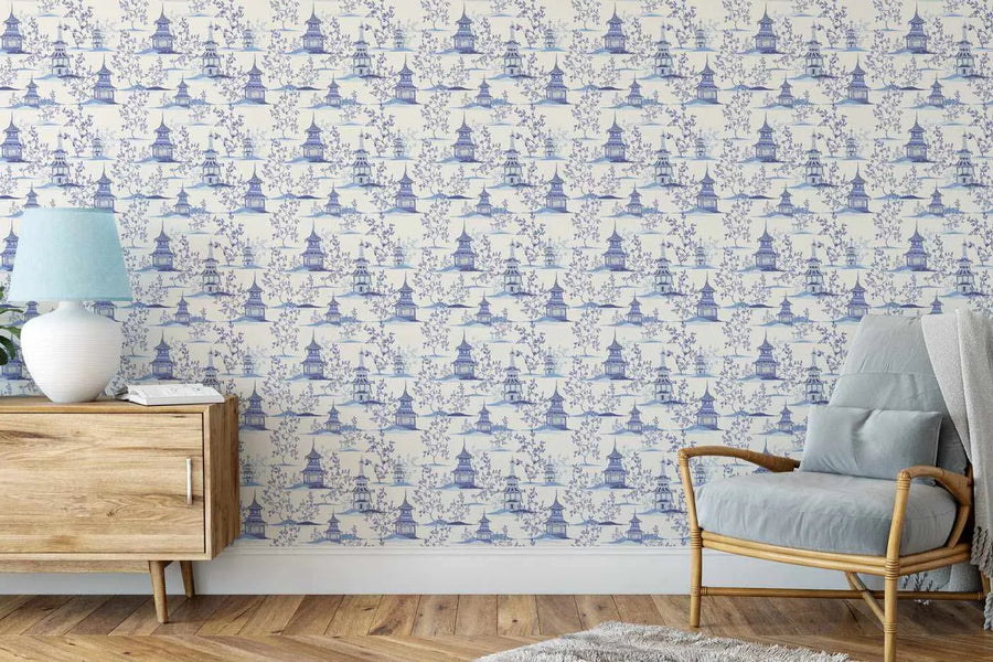Chinoiserie wallpaper featuring a repeating pattern of blue pagodas and delicate flowering trees on a cream background, displayed on a wall in a living room. The room includes a light wood sideboard with a white lamp and a light blue shade, and a rattan armchair with a light blue cushion and a grey throw.