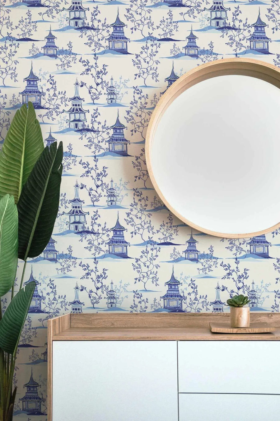 Lifestyle shot of a room featuring Chinoiserie I wallpaper with a repeating pattern of blue and white pagodas and trees. The wallpaper has a cream background with blue pagodas and trees, and a round mirror with a light wood frame is hung on the wall. A green plant is on the left side of the image, and a light wood console table with white drawers is at the bottom, with a small succulent plant in a wooden pot on top.