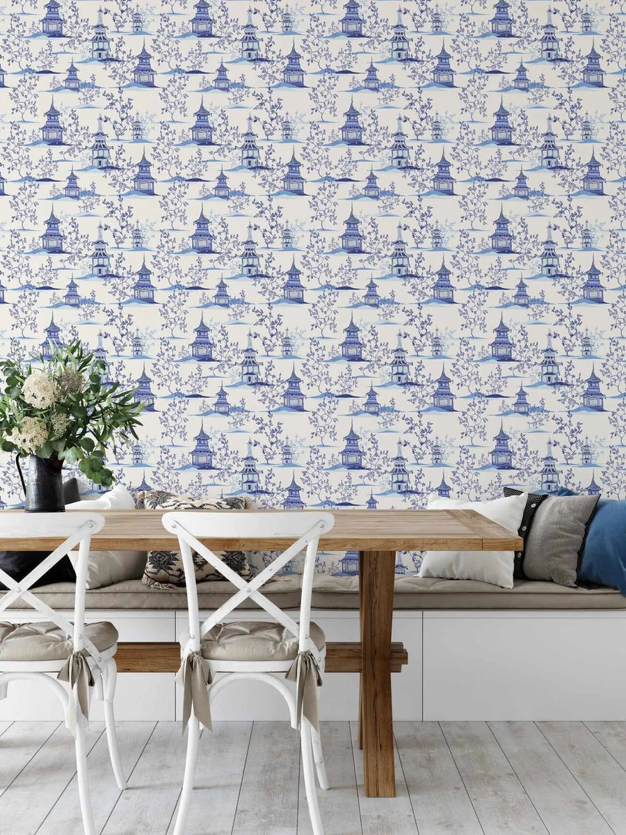 Lifestyle shot of a dining area featuring Chinoiserie I wallpaper. The wallpaper has a repeating pattern of blue and white pagodas and delicate tree branches, creating a serene, traditional Asian landscape scene. The room includes a light wood dining table, white cross-back chairs with beige seat cushions, and a built-in bench with various throw pillows in neutral and blue tones. A vase of white flowers and greenery sits on the table.