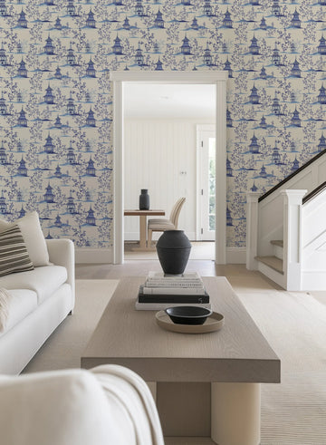 Chinoiserie I Wallpaper displayed on walls in a modern living room. The design features a repeating pattern of blue pagoda structures with ornamental flowering trees and botanical elements on a cream background. The illustration style is delicate and intricate, rendered in periwinkle blue ink. The wallpaper covers the walls flanking a white-framed doorway, creating a cohesive backdrop for minimalist furnishings including a light linen sofa, natural wood coffee table, and black ceramic vessels.