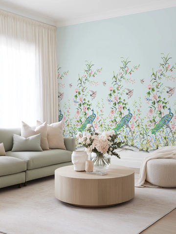 Lifestyle shot of a living room featuring Chinoiserie Luxe I wallpaper. The wallpaper has a repeating pattern of hand-painted peacocks, birds, and blooming floral branches in shades of blue, pink, and green on a pale blue background. The room includes a light green sectional sofa with cream and light green pillows, a round light wood coffee table with a vase of white and pink flowers, and a cream ottoman with a matching throw blanket.
