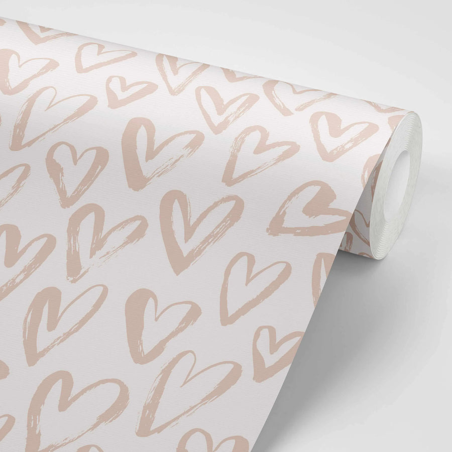 Peel and stick wallpaper featuring a repeating pattern of hand-painted hearts in a soft, muted blush pink on a white background. The hearts have a loose, brushstroke texture, giving them a hand-drawn, organic quality. The wallpaper is shown partially unrolled, revealing the pattern and the texture of the paper.