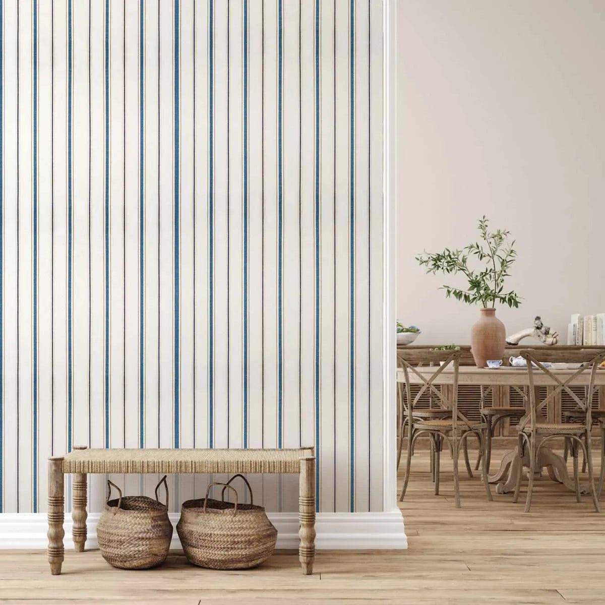 Dining Room:
Classic Nautical Stripe wallpaper featuring thin vertical stripes in deep blue and thin black lines on an off-white background, displayed on a wall in a dining room. A light wood bench with a woven top and two woven baskets underneath sits against the wallpapered wall. To the right, a dining area with a wooden table and chairs is visible, along with a vase of greenery and books on a side table.