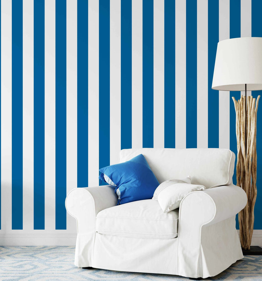 Lifestyle shot of a living room featuring Classic Stripes Wallpaper in blue and white. The wallpaper has vertical stripes of equal width in a vibrant royal blue alternating with bright white. A white slipcovered armchair with a blue cushion and a white cushion sits in the foreground, with a tall floor lamp made of natural wood branches next to it. The floor has a light blue and white patterned rug.