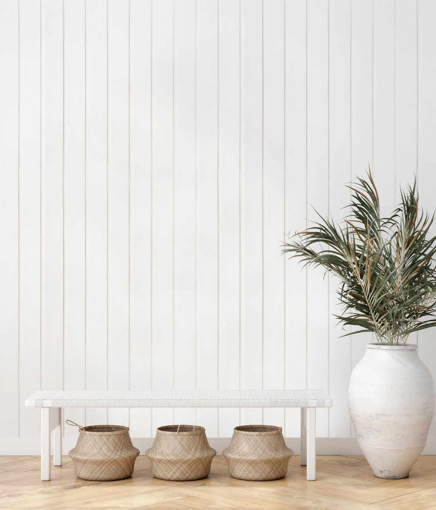 Lifestyle shot featuring Classic Tongue & Groove Wallpaper in Almost White on a wall behind a white woven bench. The wallpaper has vertical panels with subtle shadows, creating a textured, clean look. Three woven baskets sit under the bench, and a large white ceramic vase with green and brown foliage is on the right.