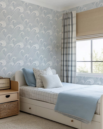 "A child's bedroom featuring a single bed with white frame and pull-out storage drawers, a light wood bedside table, and a window with a beige roman blind and a plaid curtain. The walls are adorned with a repeating pattern wallpaper of stylized ocean waves in light blue and white. The bed is dressed with a white and black grid-patterned duvet cover and a light blue textured throw blanket, with pillows in light blue, white with black speckles, and textured white."