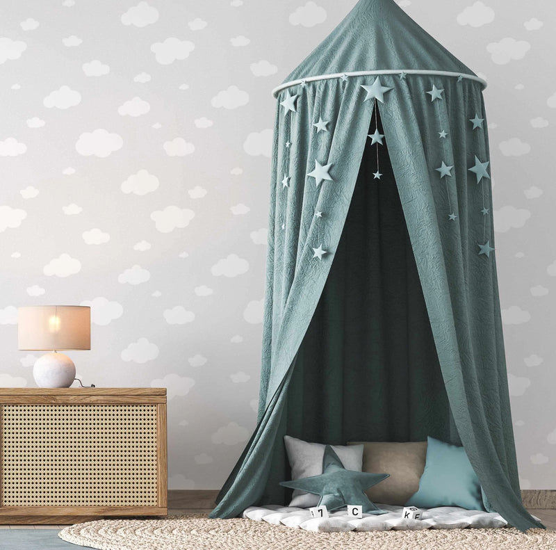 Lifestyle shot of a child's room featuring a light grey wallpaper with a repeating pattern of white clouds and subtle white polka dots. A teal canopy with hanging star decorations is set up on a woven rug, with pillows and letter blocks on the floor. A wooden side table with a lamp sits to the left.