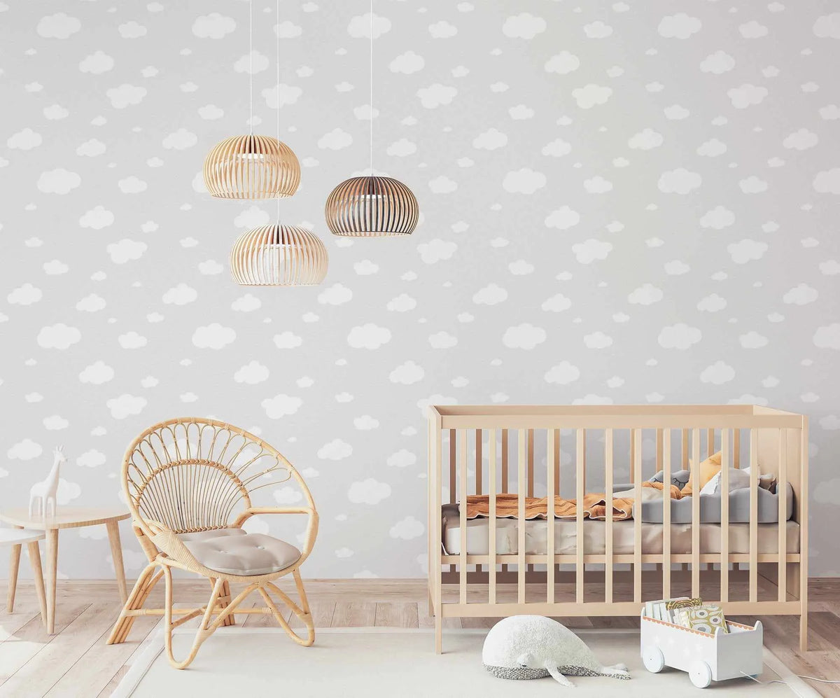 Lifestyle shot of a nursery featuring Clouds Wallpaper in Light Grey. The wallpaper has a repeating pattern of white cartoon-like clouds of various sizes on a light grey background. The room includes a light wood crib with grey and yellow bedding, a rattan chair with a light grey cushion, a small wooden side table with a white ceramic giraffe, and three wooden pendant lights hanging from the ceiling. A white whale plush toy and a white wooden toy cart are on a light-colored rug on the floor.