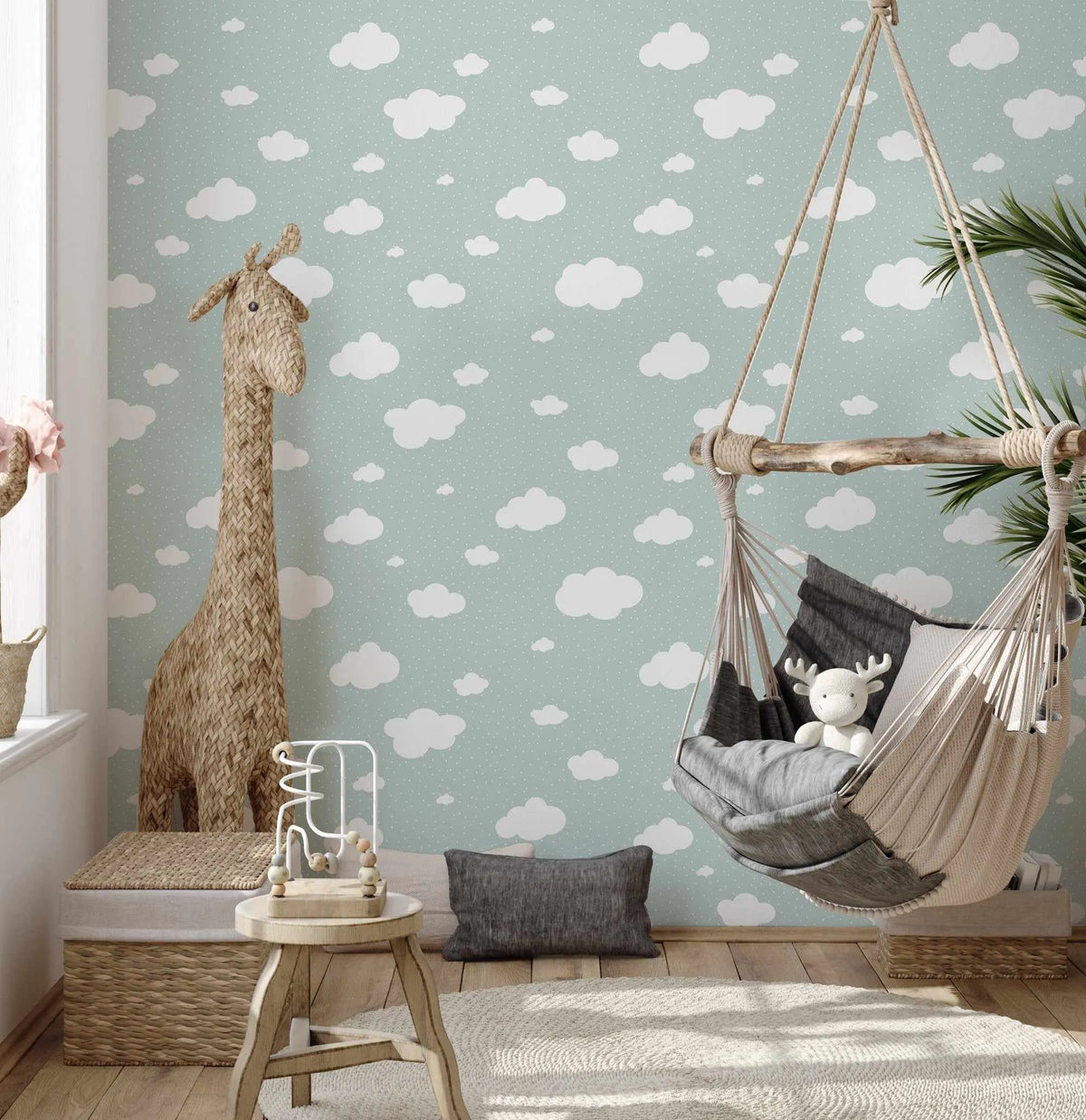 Lifestyle shot of a child's room featuring Clouds Wallpaper in Sage. The wallpaper has a repeating pattern of fluffy white clouds of varying sizes and small white polka dots on a soft sage green background. A large woven giraffe stands in the left corner, and a natural wood and rope hammock chair hangs from the ceiling on the right, with a white stuffed reindeer toy inside. The room has light wood flooring, a cream textured rug, and a small wooden stool with a white and wood bead maze toy.