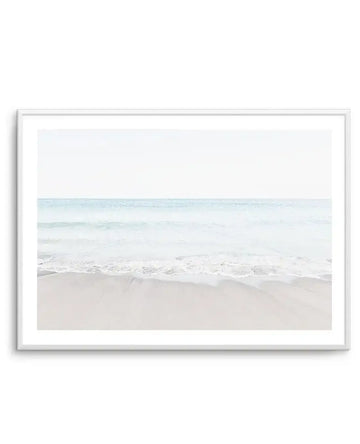 Framed photography art print of a serene coastal beach scene with a white frame and white border. The composition features gentle, light blue waves breaking on a wide expanse of pale, sandy beach under a bright, almost white sky.