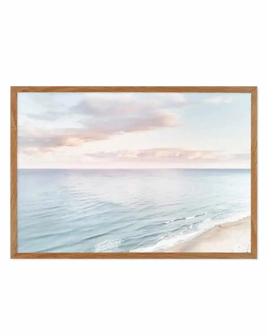 Framed photography art print of a coastal sunset scene with natural wood frame and white border. The composition features a wide expanse of calm ocean with gentle waves breaking on a sandy beach, under a sky with soft pink and blue clouds.