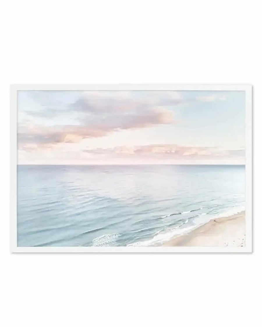 Framed photograph of a coastal beach scene at sunset with a white frame and white border. The composition features gentle waves rolling onto a sandy beach under a soft sky with pastel pink and blue clouds, creating a serene and expansive mood.