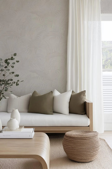Coastal Luxe Palms wallpaper in warm grey displayed on a living room wall behind a light wood sofa. The wallpaper features a subtle, tonal palm frond pattern in soft grey and beige tones creating a layered, textured appearance. The room shows the wallpaper's understated botanical design paired with cream linen cushions in coordinating neutral tones, white curtains, a woven rope ottoman, and natural wood furnishings, demonstrating the product's calming, coastal aesthetic in an interior setting.