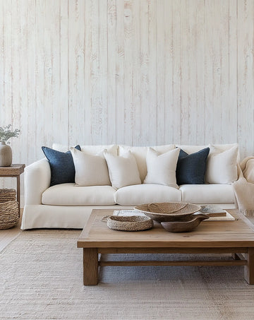 "Coastal whitewashed wood panels wallpaper installed on a wall behind a white sofa in a bright living room. The wallpaper features vertical planks with visible wood grain and knots in shades of off-white and pale beige, creating a rustic yet airy backdrop. The room includes a wooden coffee table with woven bowls, a jute rug, and throw pillows in cream and deep blue."