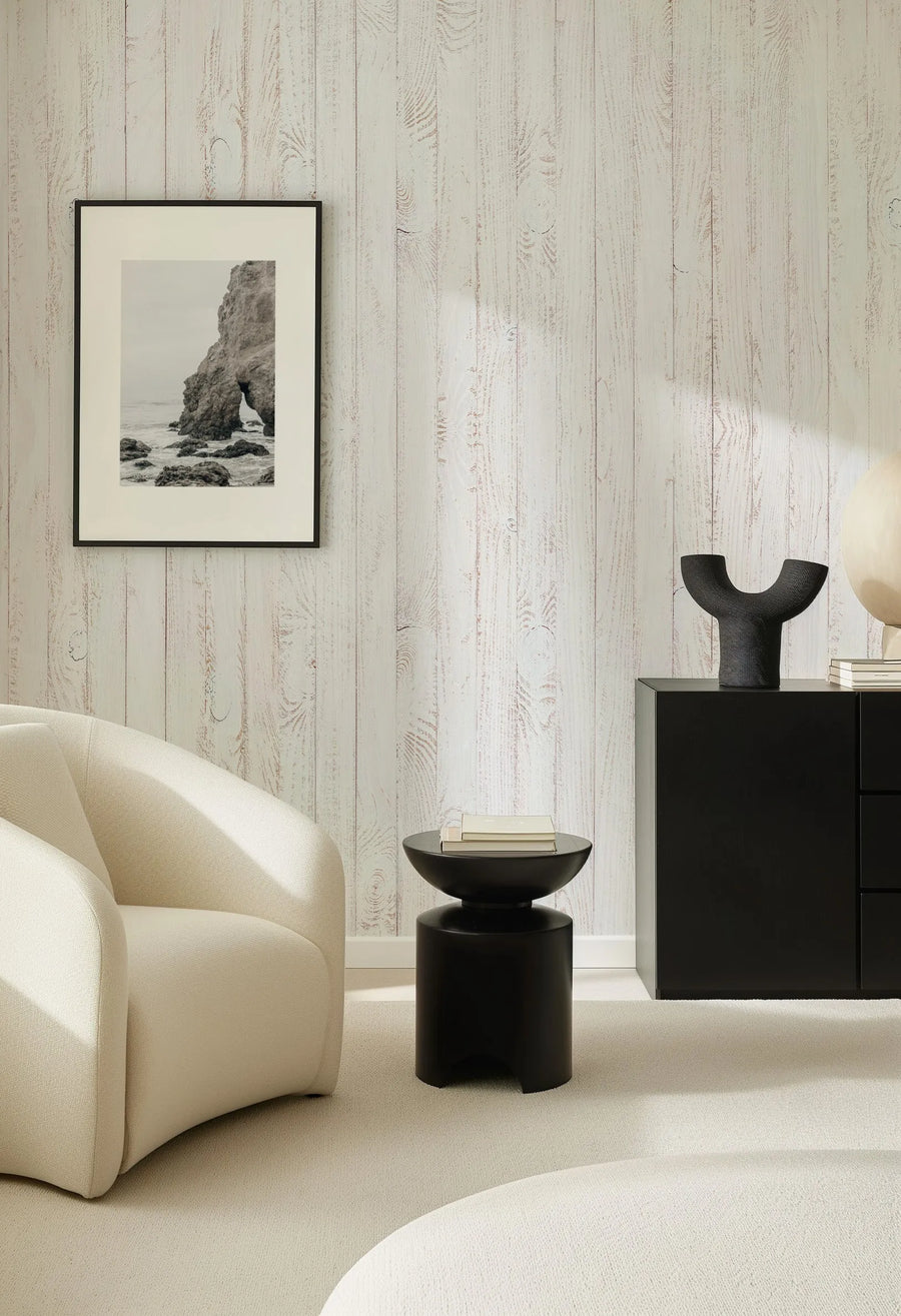 Lifestyle shot of a minimalist living room featuring Coastal Whitewashed Wood Panels Wallpaper. The wallpaper has a repeating pattern of vertical whitewashed wooden planks with visible wood grain and subtle reddish-brown undertones, creating a rustic yet clean aesthetic. A framed black and white photograph of a rocky coastal scene hangs on the wall. A curved cream-coloured armchair sits in the foreground, and a black side table with books and a black cabinet with decorative objects are to the right.