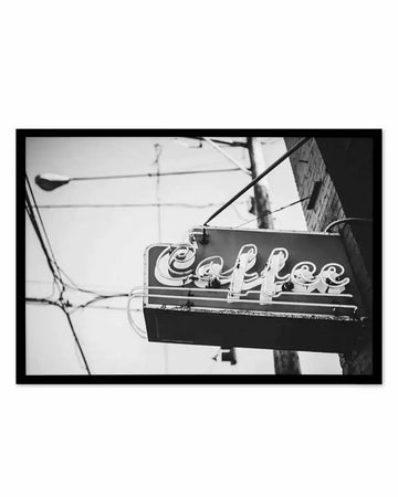 Framed black and white photograph of a vintage "Coffee" neon sign, captured from a low angle against a bright, overcast sky. The sign, with its cursive lettering, hangs from a pole, with electrical wires and a streetlamp visible in the background, conveying an urban, nostalgic mood.