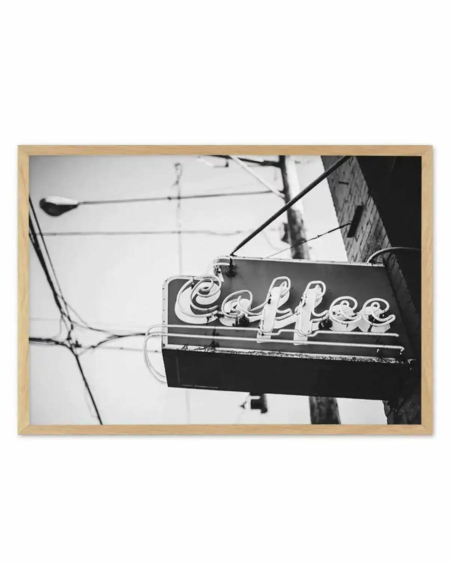 Framed black and white photograph of a vintage "Coffee" neon sign hanging outdoors. The sign features stylized, cursive lettering with a double outline, suggesting neon tubes, against a dark rectangular background. It is attached to a brick building and supported by metal poles and wires, with a street lamp and more utility wires visible in the soft-focus background, all displayed in a natural oak frame with white border.