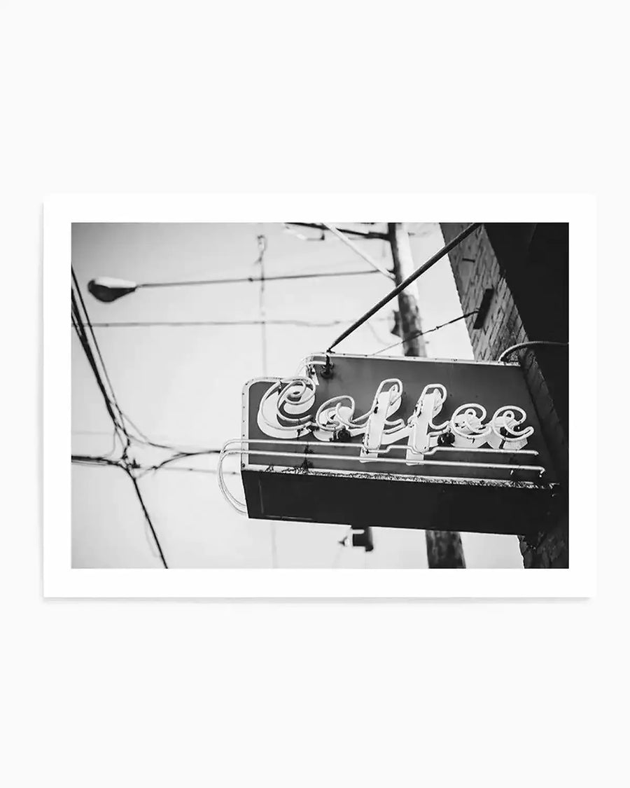Framed black and white photograph of a vintage neon "Coffee" sign hanging from a brick building, with a street lamp and power lines visible in the background against a bright sky. The sign features stylized script lettering with visible neon tubes, and the overall composition has a gritty, urban feel.