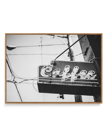 Black and white photograph of a vintage neon "Coffee" sign mounted on a dark rectangular housing, with power lines and a brick building visible in the soft-focused background. The neon lettering glows against the dark sign face. The photograph is printed on canvas and housed in a natural wood frame with a white border.