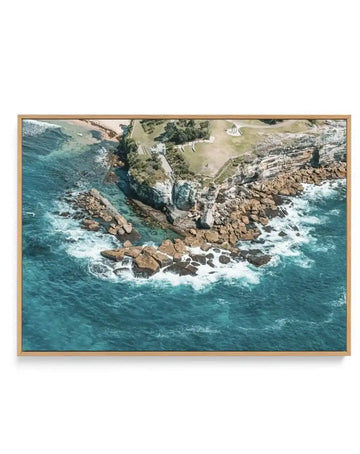 "Aerial photograph printed on canvas, depicting a coastal scene with a rocky shoreline, turquoise ocean, and a grassy headland. The composition features waves crashing against brown rocks in the foreground, leading to a small sandy beach and a green park area with structures on the cliff edge. The canvas is displayed in a natural wood frame."