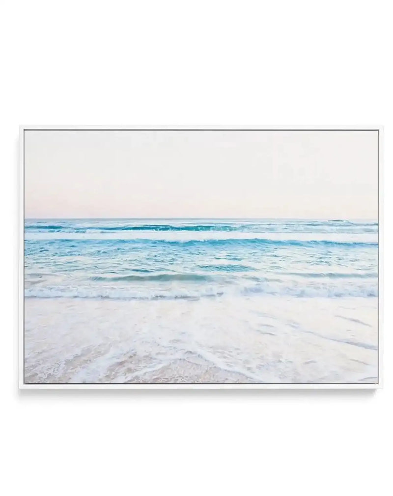 Framed canvas print of a serene coastal beach scene with a white frame. The photograph captures gentle, light blue waves breaking on a wide expanse of white sand, with a soft, pale sky stretching to the horizon.