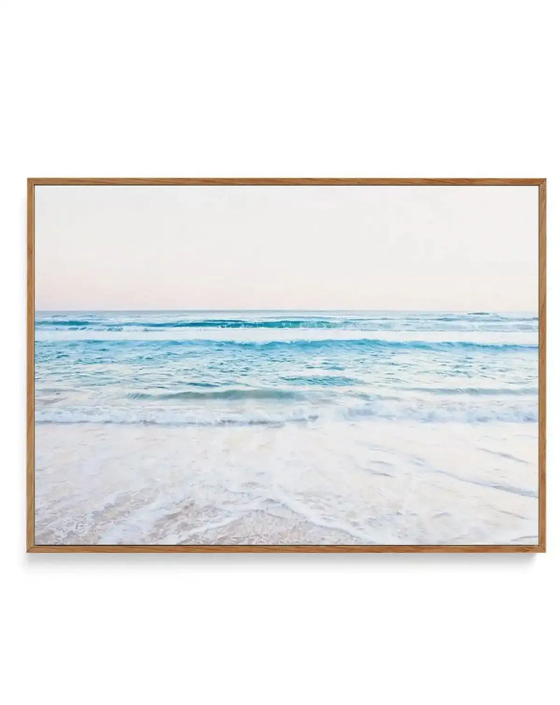 Framed canvas print of a serene coastal beach scene with a natural wood frame. The photograph captures gentle, light blue waves breaking on a white sandy shore, with a soft, pale sky stretching to the horizon.