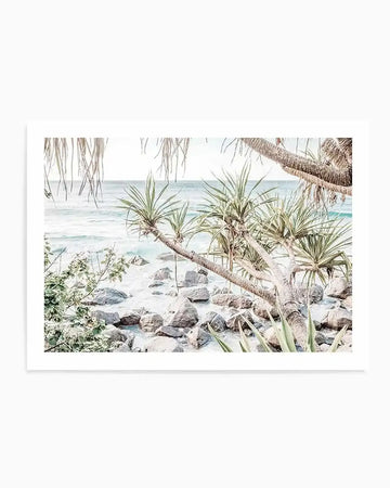 Framed photography art print of a coastal scene with a natural wood frame and white border. The composition features a sandy beach with scattered rocks and gentle turquoise waves, viewed through the tropical foliage of pandanus trees with long, slender leaves and visible root systems.