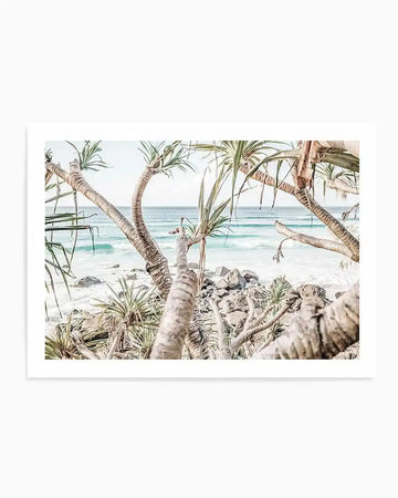 Coastal landscape art print featuring gnarled palm tree trunks in foreground with green fronds, framing a turquoise ocean view with white sandy beach and scattered rocks. Soft, muted colour palette of cream, sage green, and pale blue creates a serene tropical scene. Watercolour illustration style with delicate detail. Mounted on white background with white border.