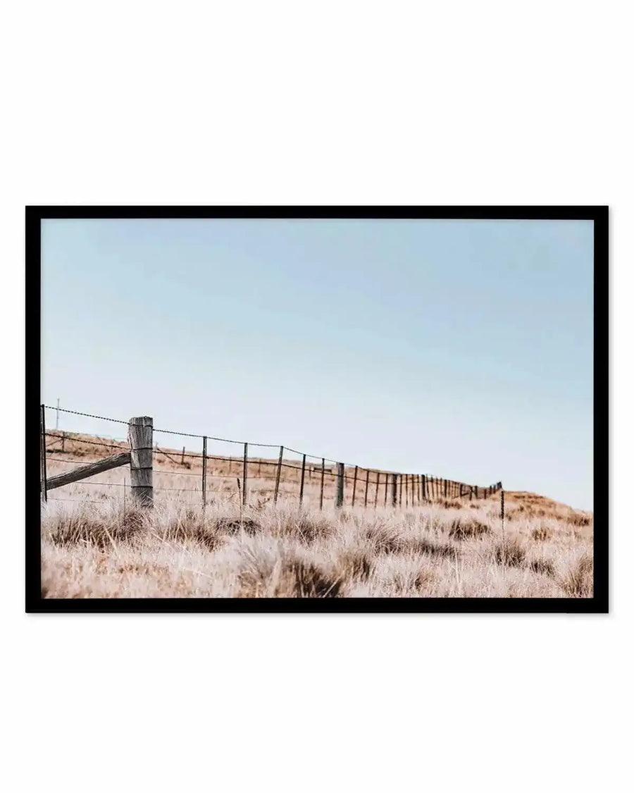 Framed photography art print featuring a countryside scene with a wooden fence line stretching across a dry, grassy hill under a pale blue sky, displayed in a black frame with white border.