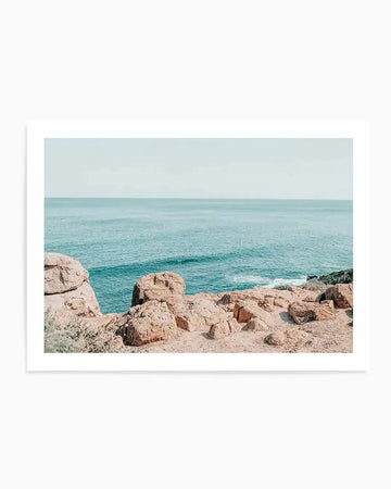 Framed photography art print of a coastal scene with a natural wood frame and white border. The composition features a calm ocean with gentle waves under a pale blue sky, with a foreground of reddish-brown rocks and sandy ground.