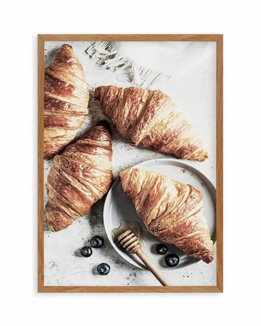 Framed photography art print of a breakfast scene with four golden-brown croissants, a honey dipper with honey, and blueberries on a light grey surface, displayed in a natural oak frame with white border. Three croissants are arranged on the surface, and one is on a white plate.