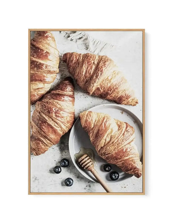 Framed canvas art print featuring a flat lay food photography composition of four golden-brown croissants with flaky, layered texture arranged on a white speckled surface. One croissant rests on a white ceramic plate with a honey dipper beside it. Fresh blueberries are scattered around the arrangement. The print is mounted in a natural wood frame with white border.